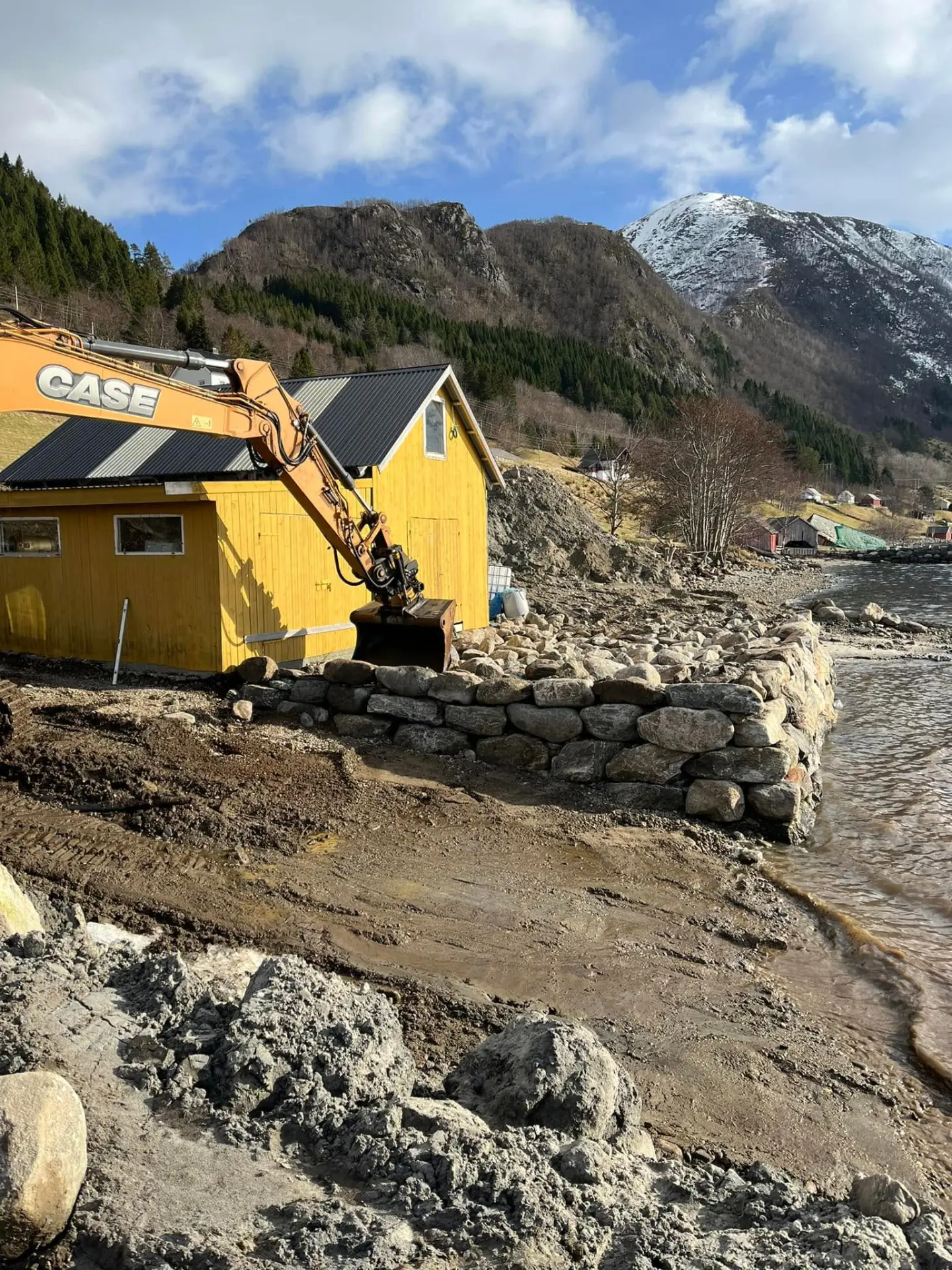 Gravemaskin bygger sjømur i stein ved gult naust