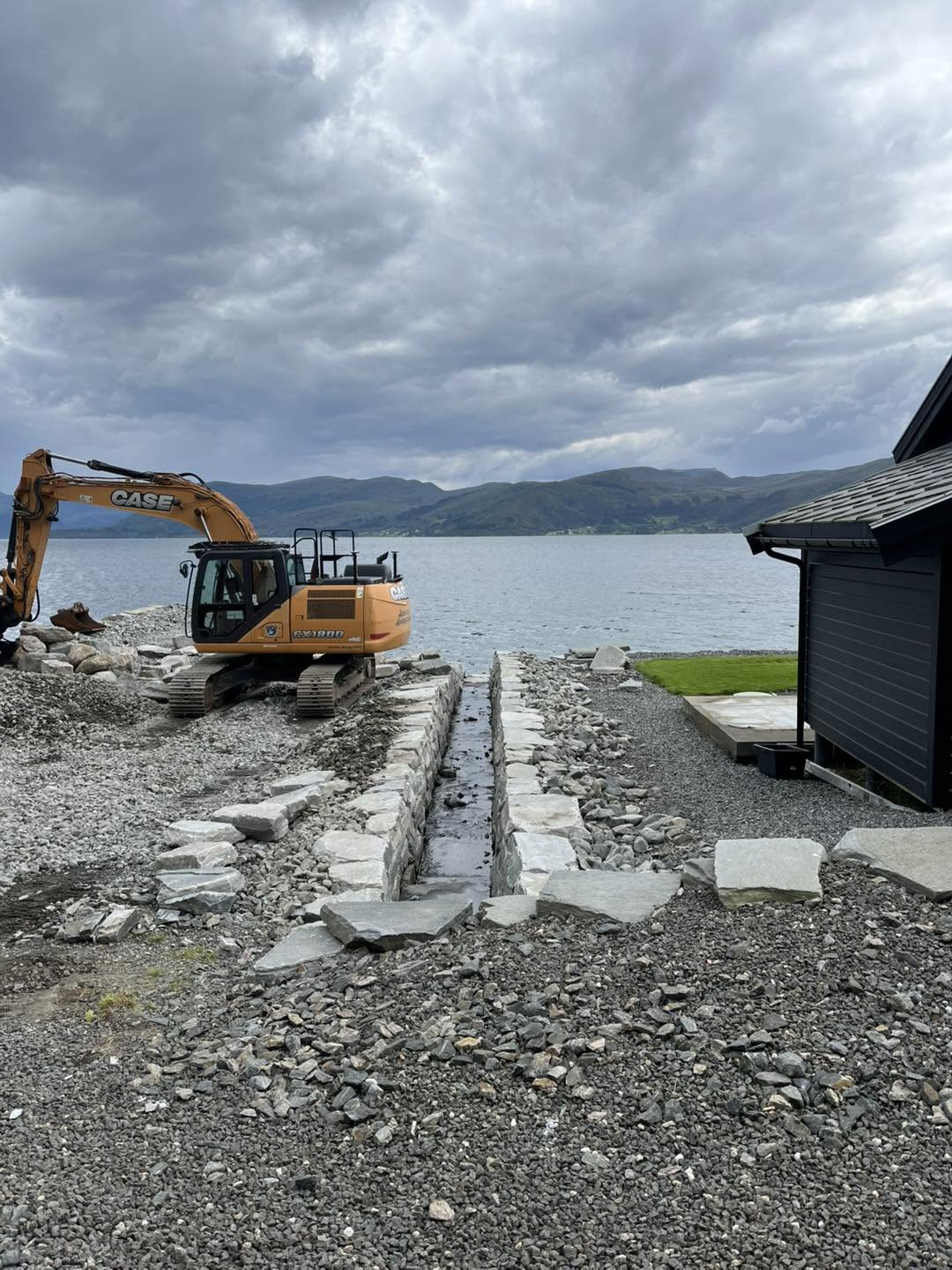 Naturstein lagt for hand ved steinrenne ned mot fjorden, med gravemaskin i bakgrunnen