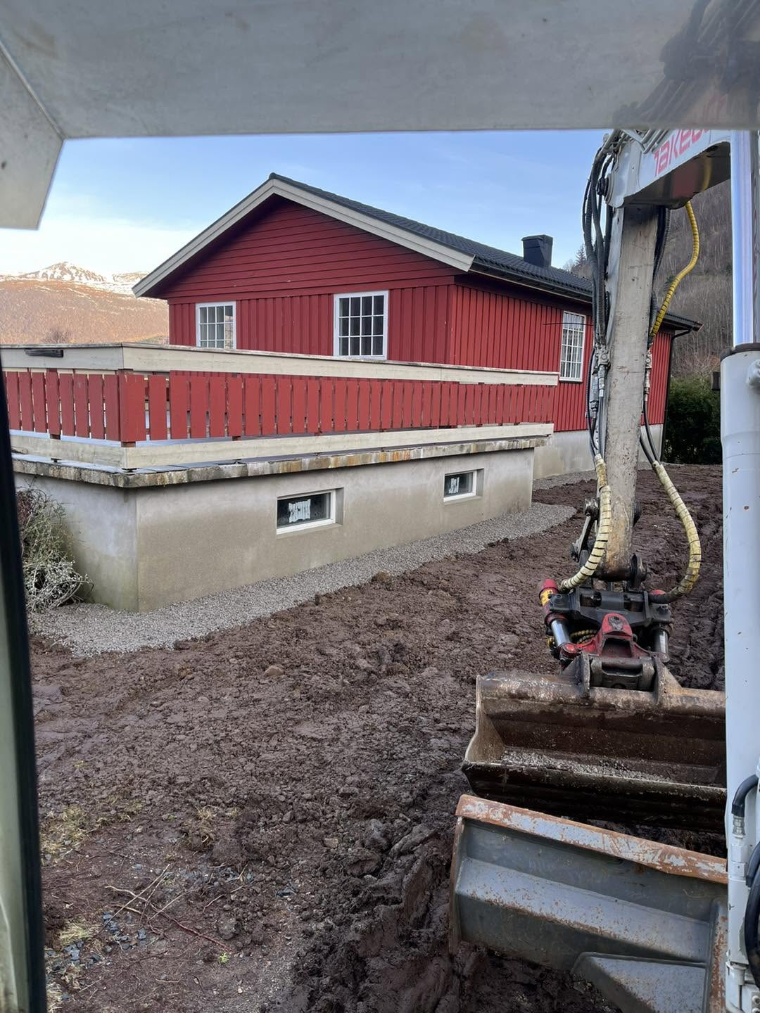 Gravemaskin planerer gjørmete uteareal rundt raudt trehus